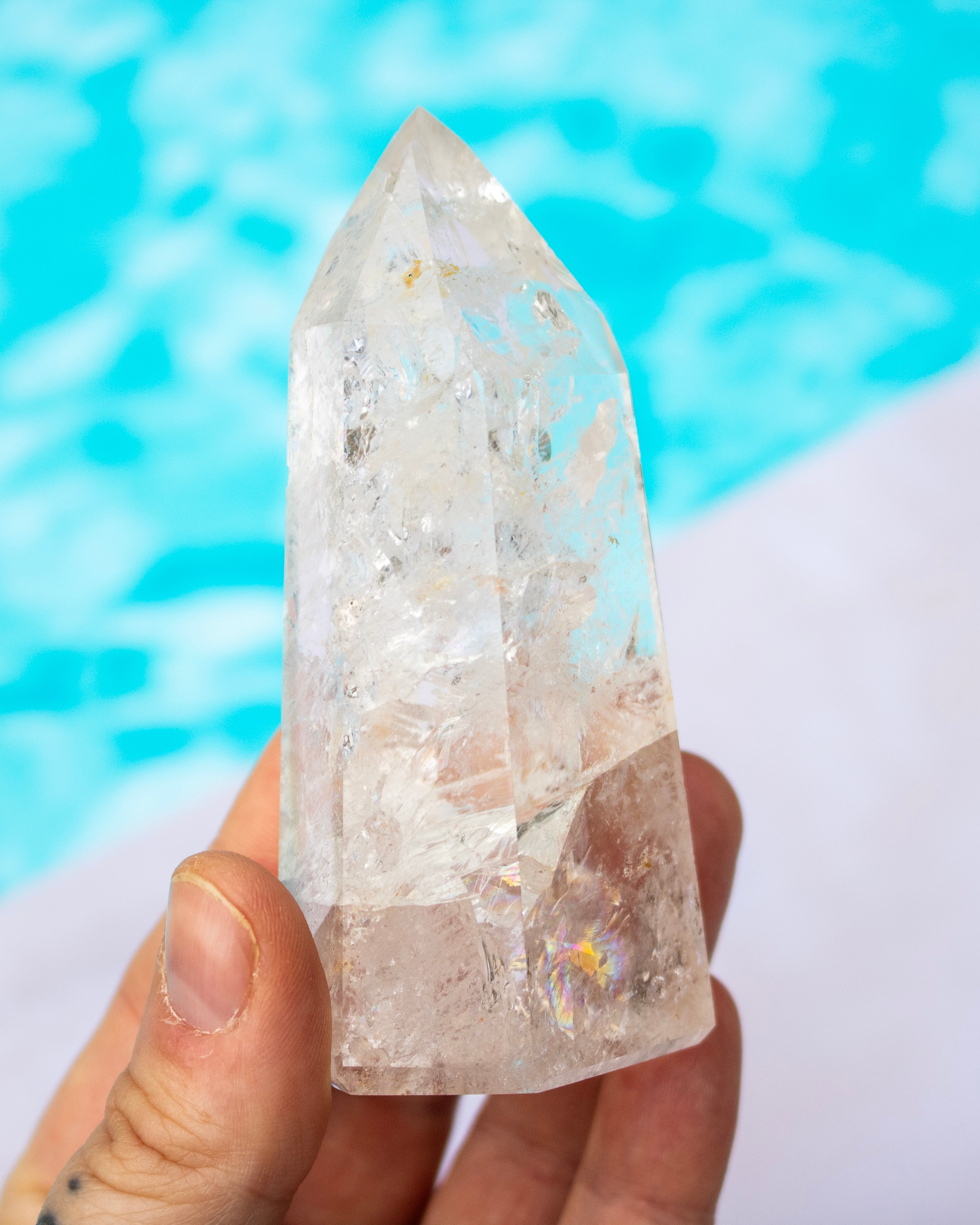 A hand holding a clear quartz crystal point with natural inclusions, set against a blurred background of bright blue water and white sand.