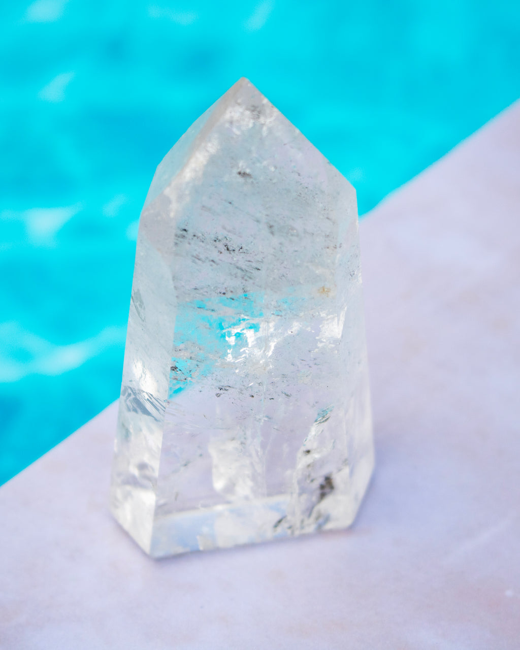 A clear pointed crystal tower or quartz point with natural internal inclusions, standing on a pale surface with a bright turquoise blue blurred background.