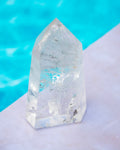 A clear pointed crystal tower or quartz point with natural internal inclusions, standing on a pale surface with a bright turquoise blue blurred background.