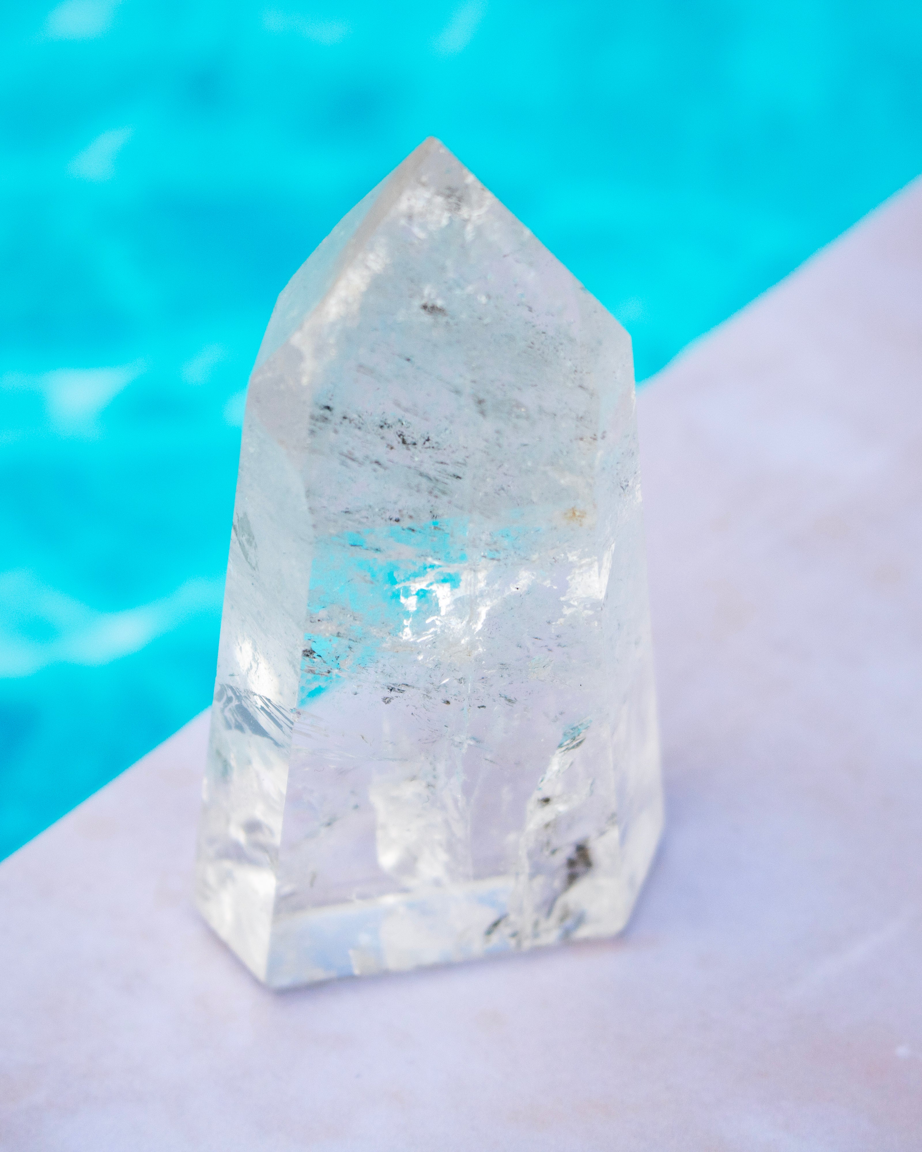 A clear pointed crystal tower or quartz point with natural internal inclusions, standing on a pale surface with a bright turquoise blue blurred background.