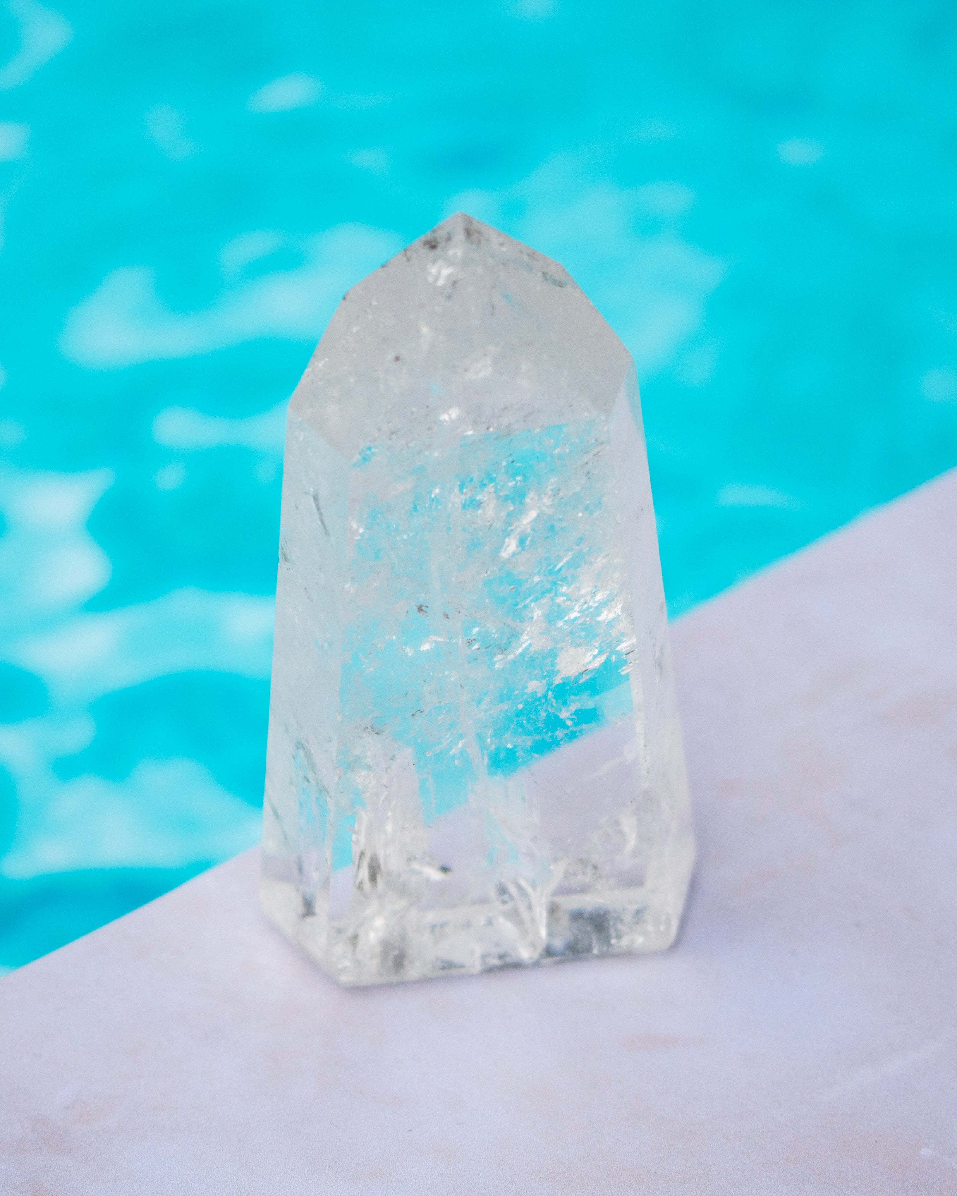 A clear natural quartz crystal tower with internal inclusions, resting on a white stone ledge with a bright turquoise pool in the background.