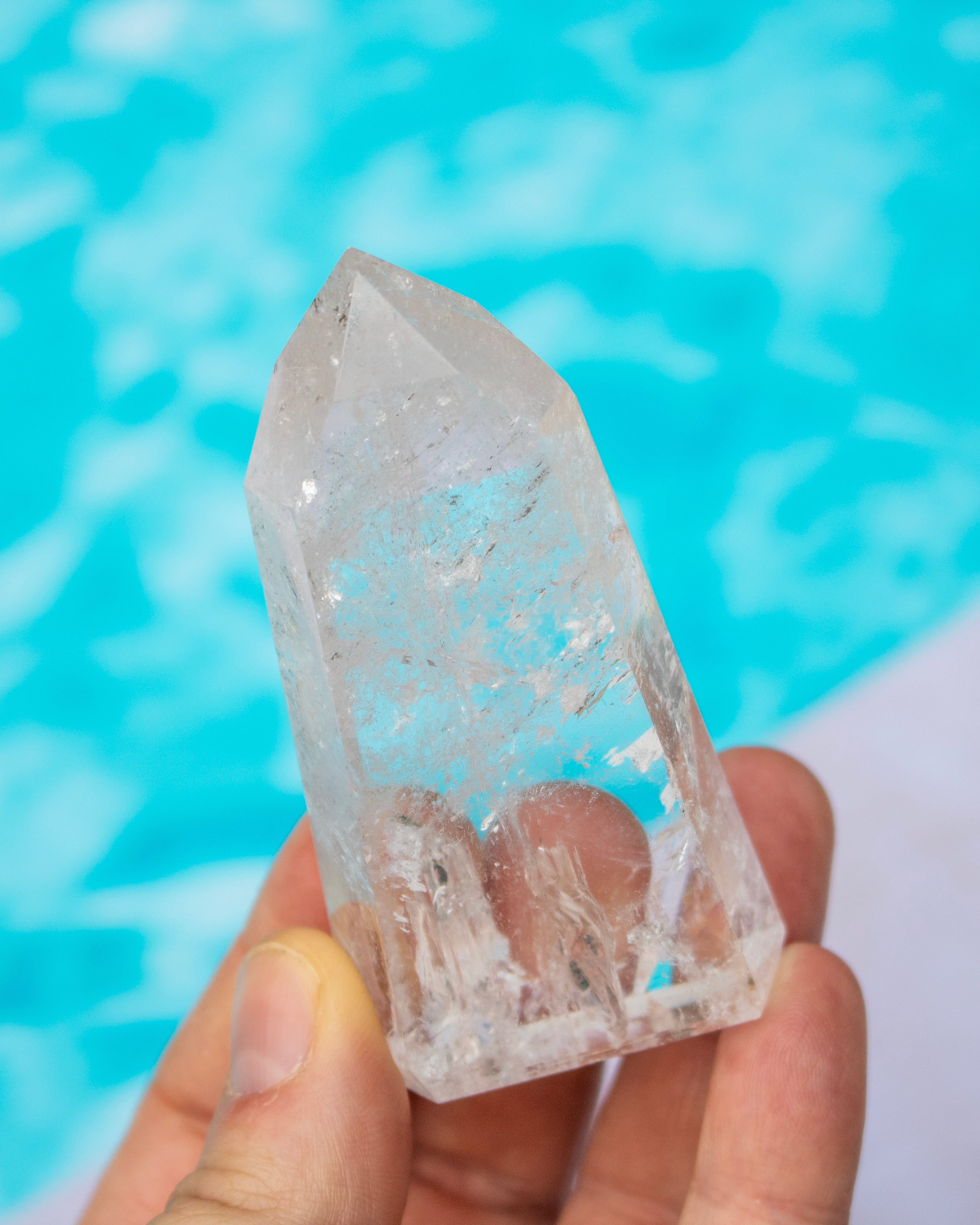A clear quartz crystal point held in a person's hand, with a bright turquoise pool background.