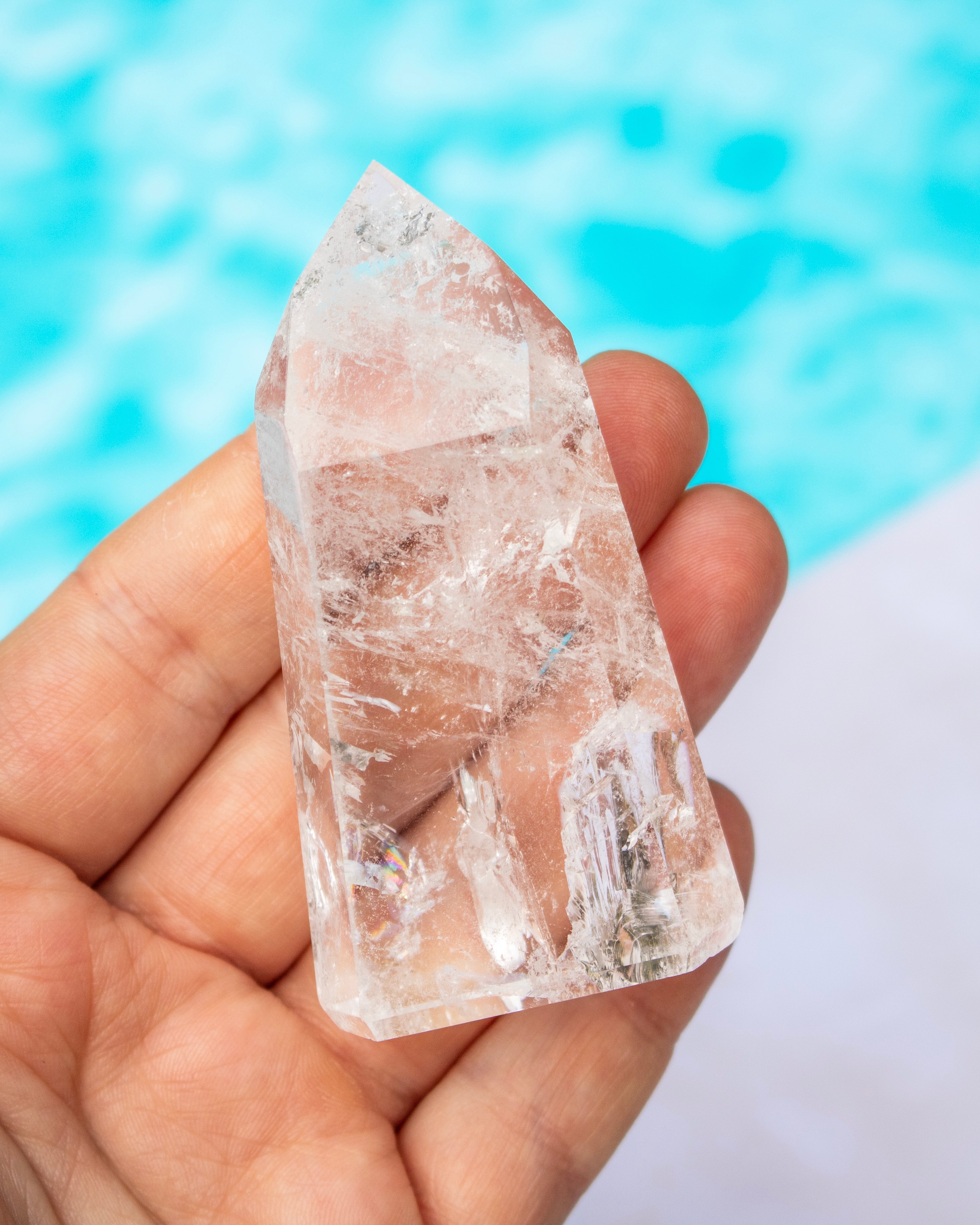 A hand holding a pointed clear quartz crystal against a blurred background of bright blue water and white sand.