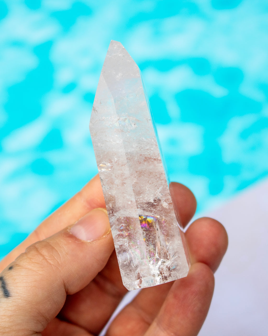 A hand holding a clear pointed crystal tower with natural internal inclusions, set against a bright turquoise blue blurred background.