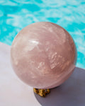 A large polished rose quartz sphere resting on a small gold-colored stand, set against a blurred background of a swimming pool.