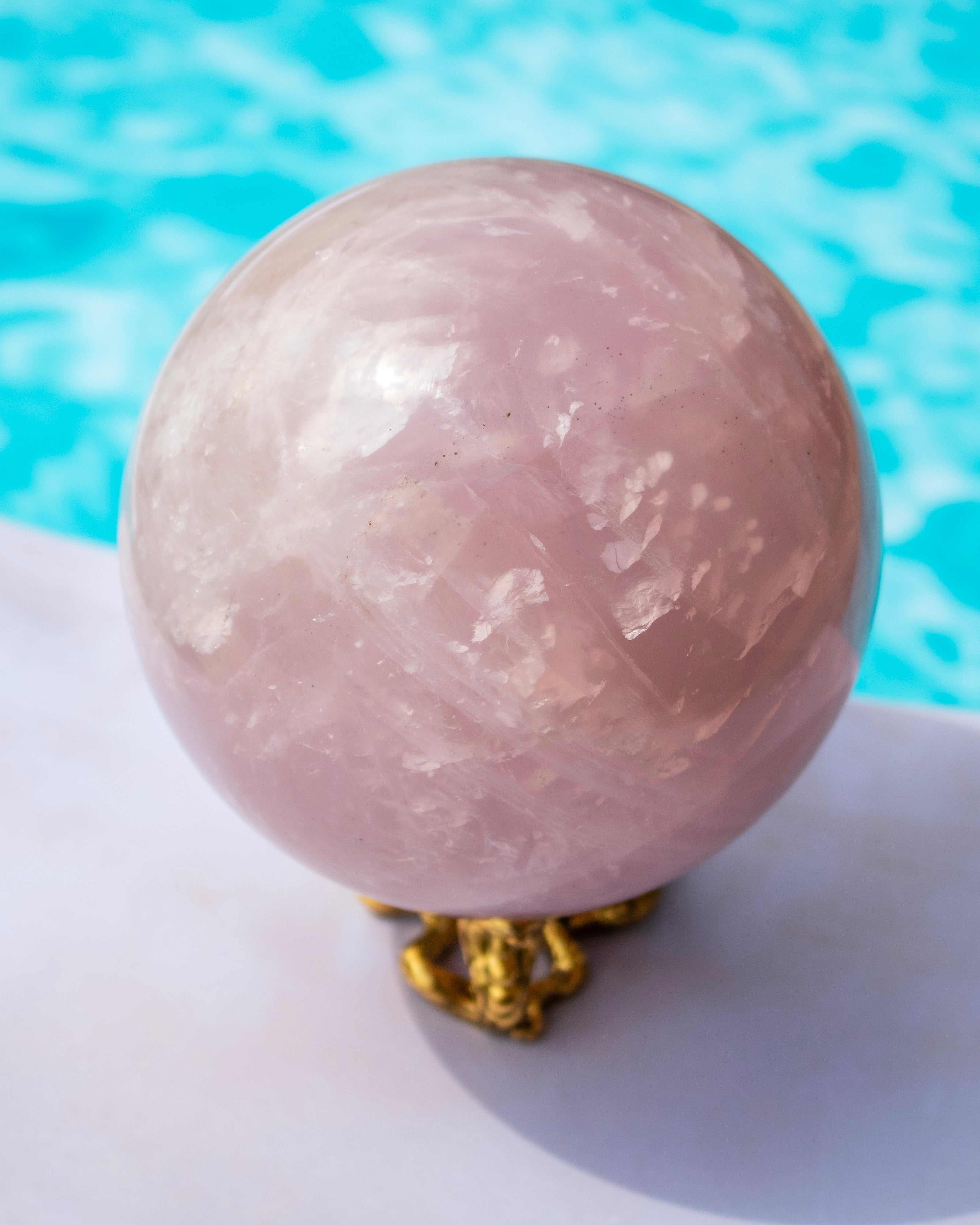 A polished pink rose quartz crystal sphere resting on a small gold-colored metal stand, set against a blurred background of bright blue water.