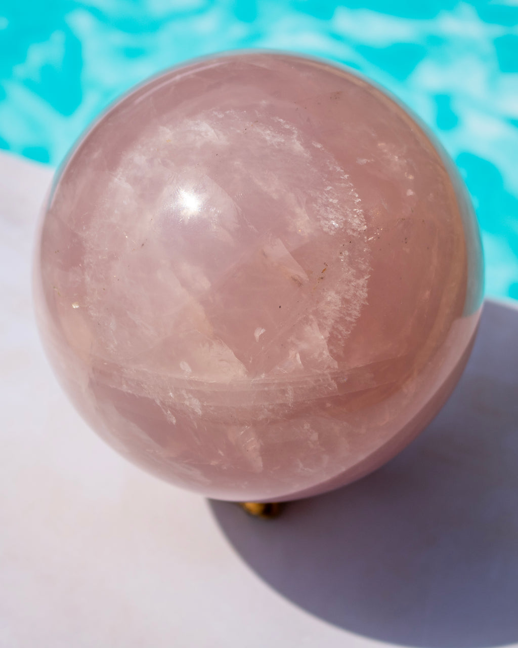 A large polished rose quartz crystal sphere resting on a white surface with a bright turquoise blue background.