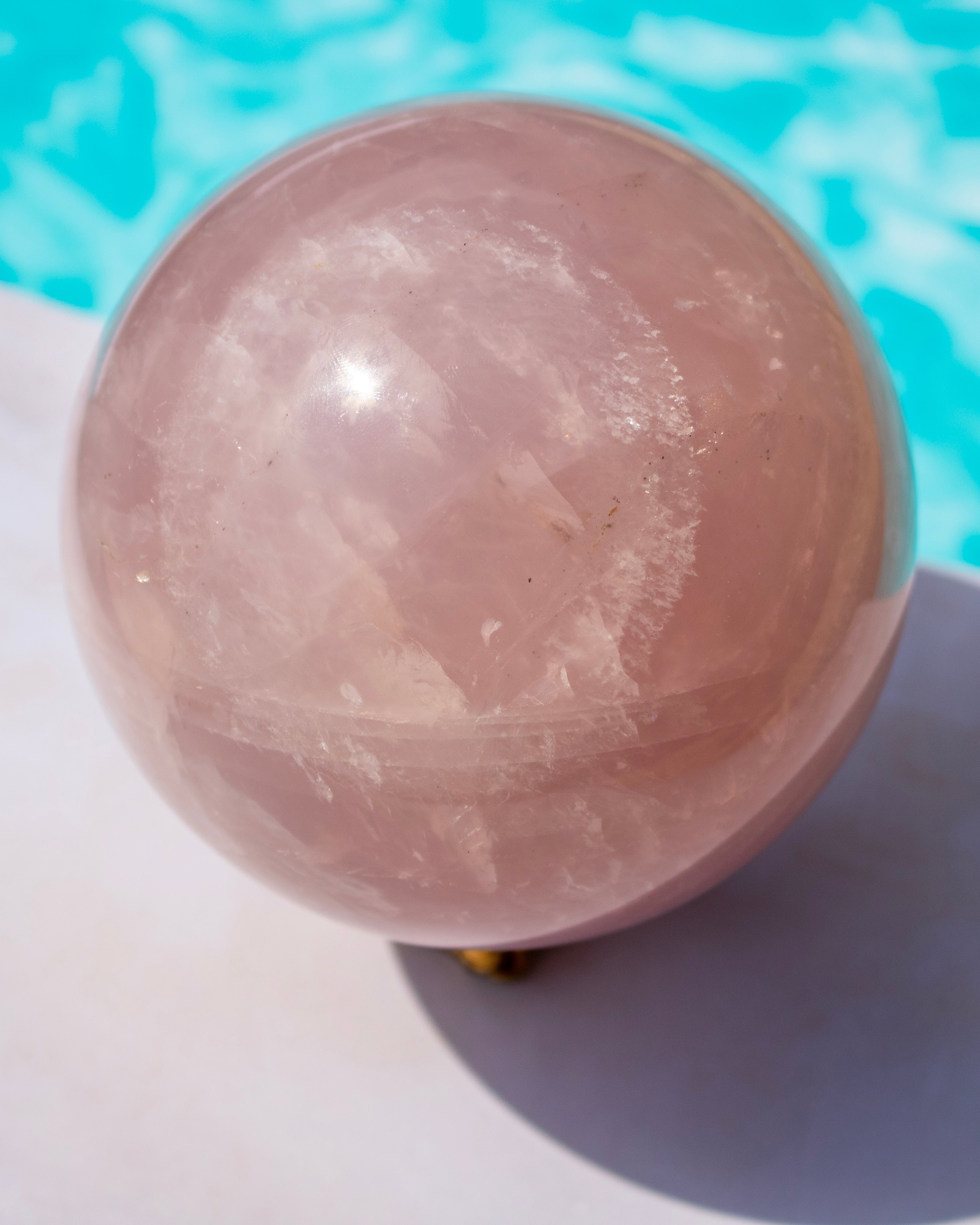 A large polished rose quartz crystal sphere resting on a white surface with a bright turquoise blue background.
