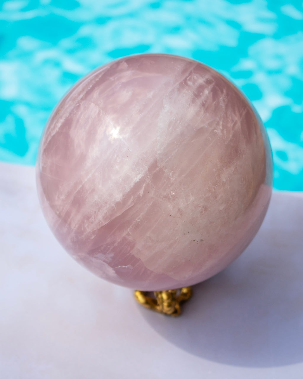 A polished pink quartz crystal sphere resting on a small gold stand, set against a bright turquoise pool background.
