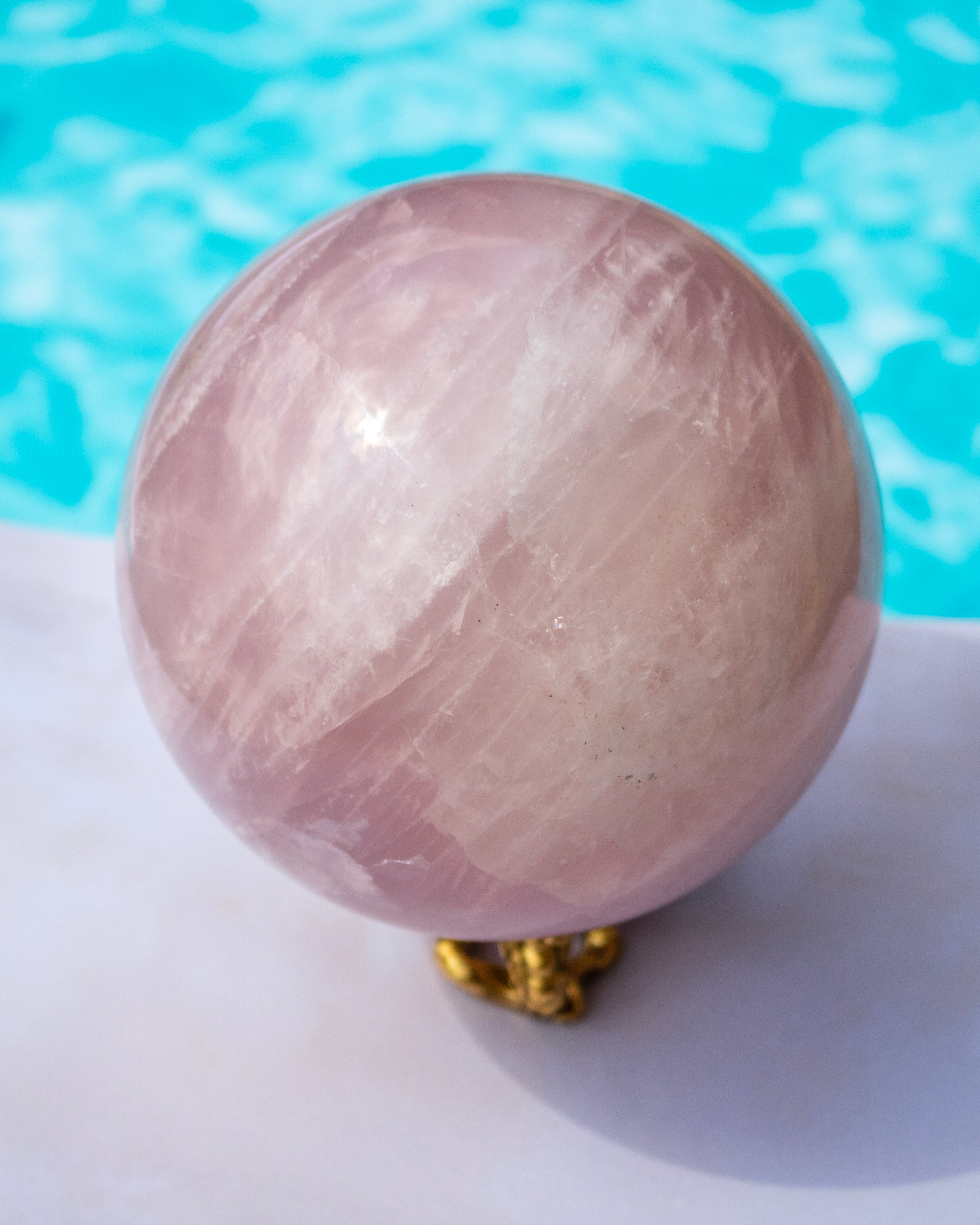 A polished pink quartz crystal sphere resting on a small gold stand, set against a bright turquoise pool background.