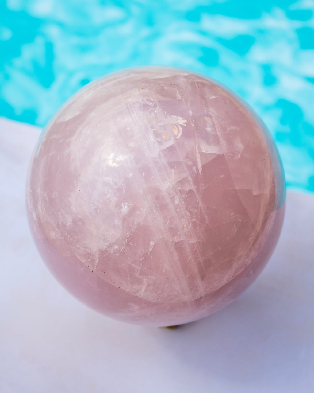 A polished pink rose quartz crystal sphere resting on a white surface with a blurred turquoise blue background.