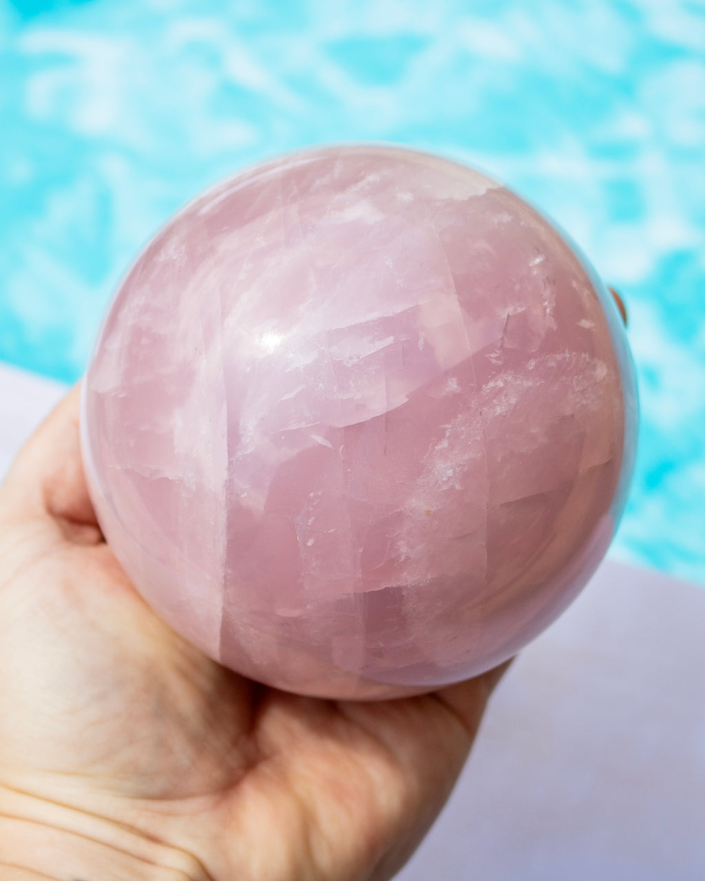 A hand holding a polished pink rose quartz crystal sphere, with a blurred light blue background.