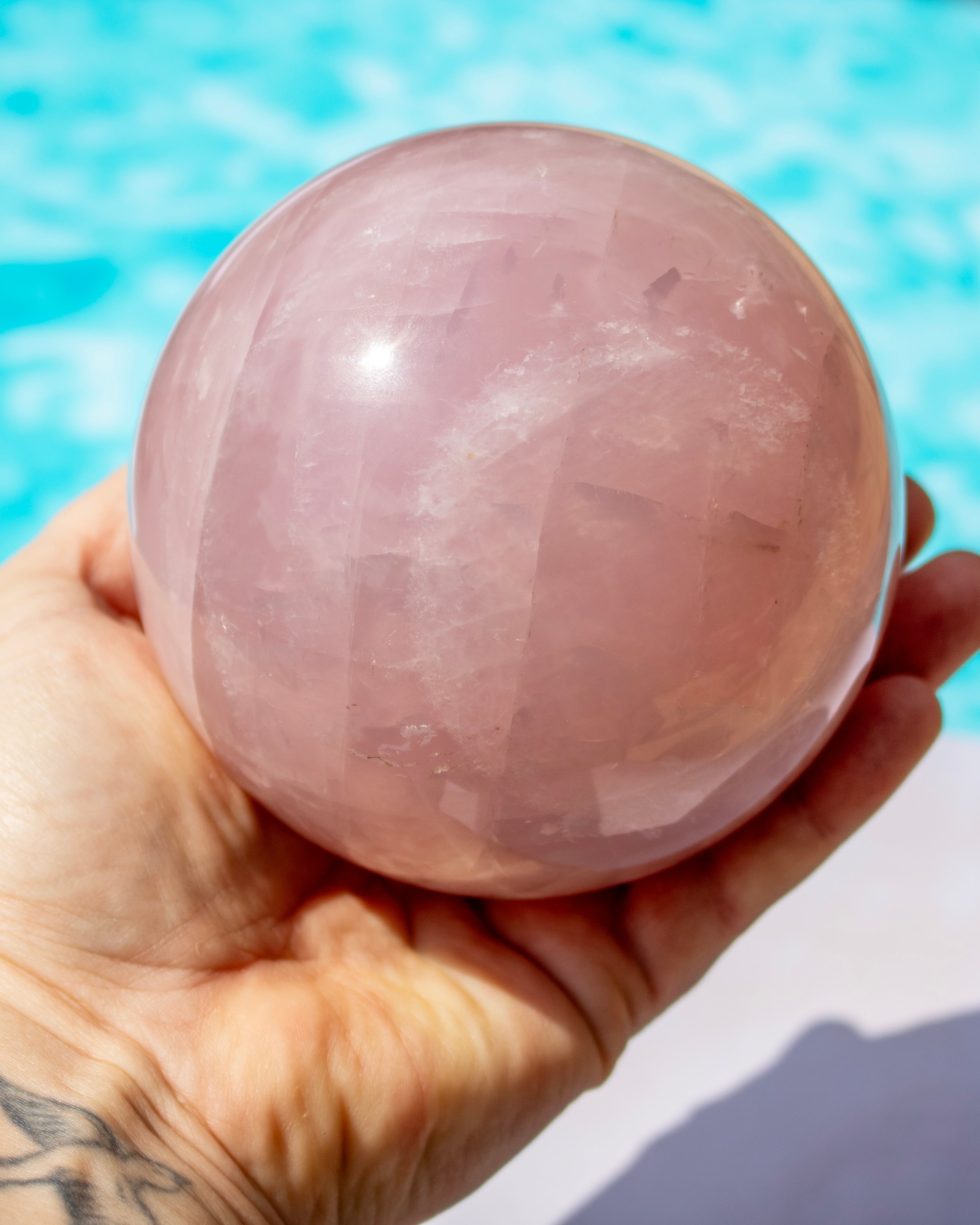 A hand holding a large, polished pink rose quartz crystal sphere, with a blurred turquoise swimming pool in the background.