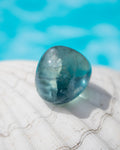 A polished teardrop-shaped blue gemstone resting on a white seashell with a blurred turquoise water background.