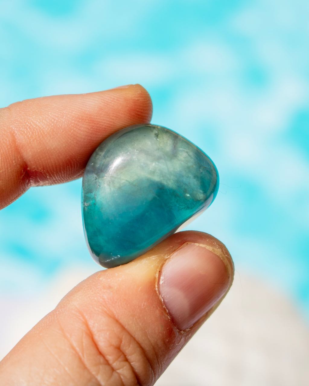 A polished tumbled blue stone held between two fingers against a bright, blurred turquoise background.
