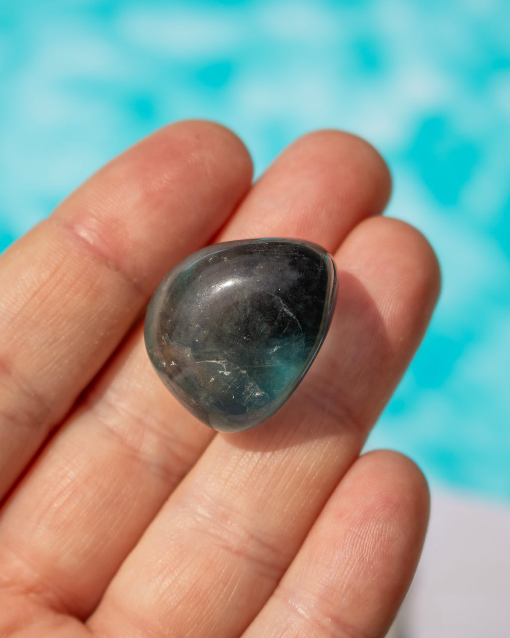 A polished dark gray tumbled stone held in a person's hand, with a bright turquoise-blue blurred background.