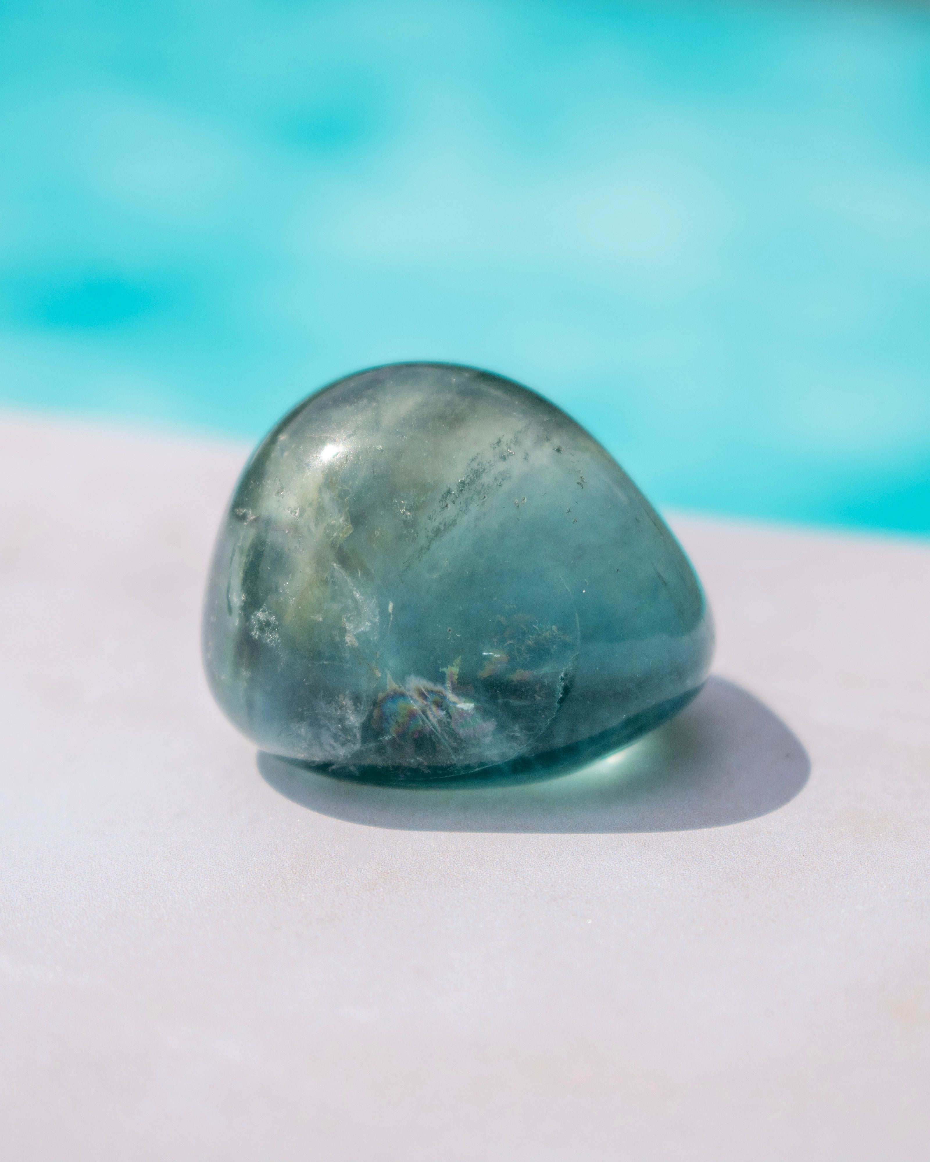 A polished tumbled stone in a muted teal-blue color, resting on a light surface with a soft-focus turquoise background.
