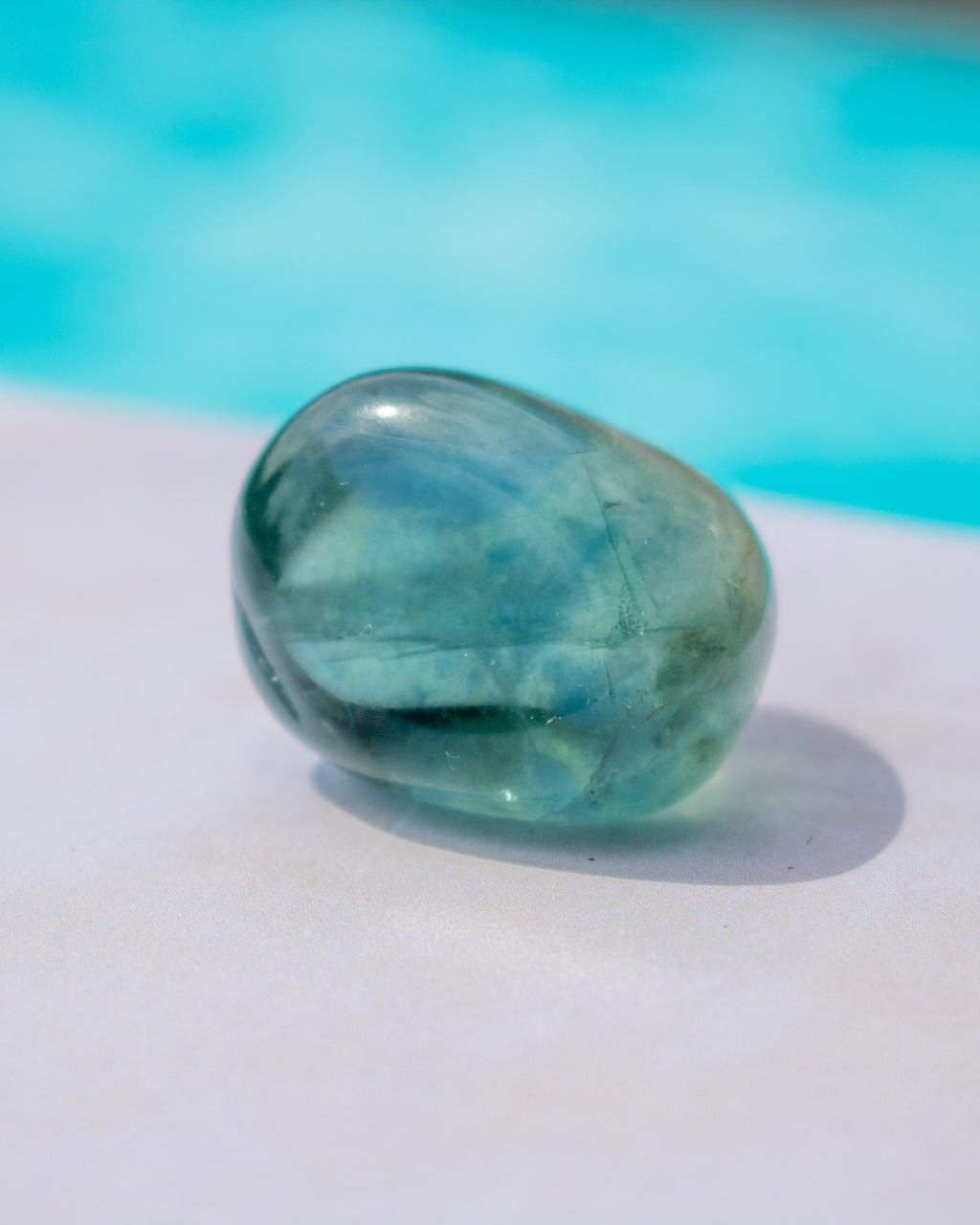 A polished tumbled aquamarine stone, showing a translucent light blue-green color, resting on a white surface with a blurred turquoise background.