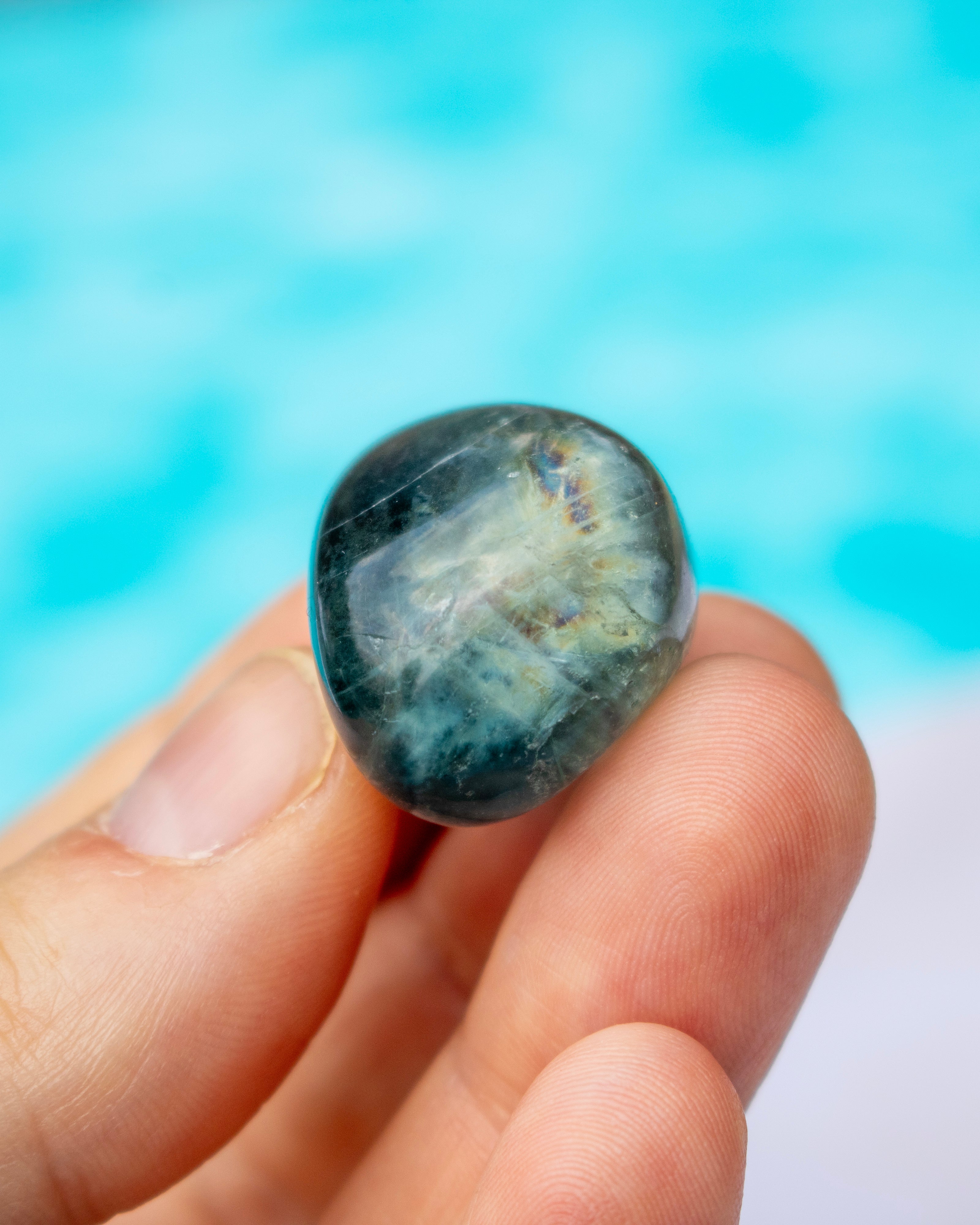 A hand holding a polished, teardrop-shaped dark blue gemstone with white and gray crystalline patterns, set against a bright, blurred turquoise background.