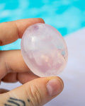 A hand holding a smooth, oval-shaped translucent pink stone or crystal against a blurred turquoise and white background.