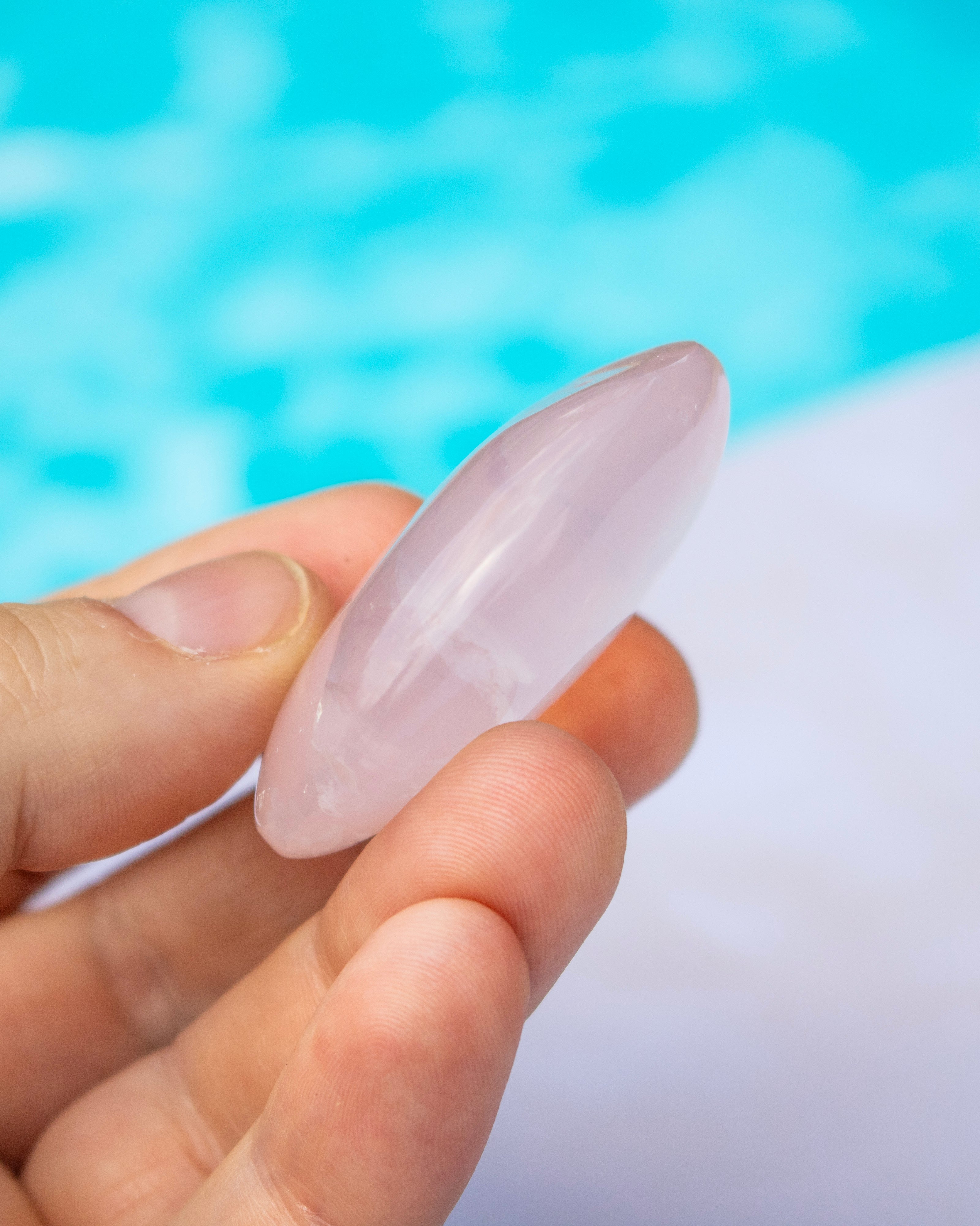 A hand holding a smooth, polished pale pink stone or crystal against a blurred turquoise and white background.