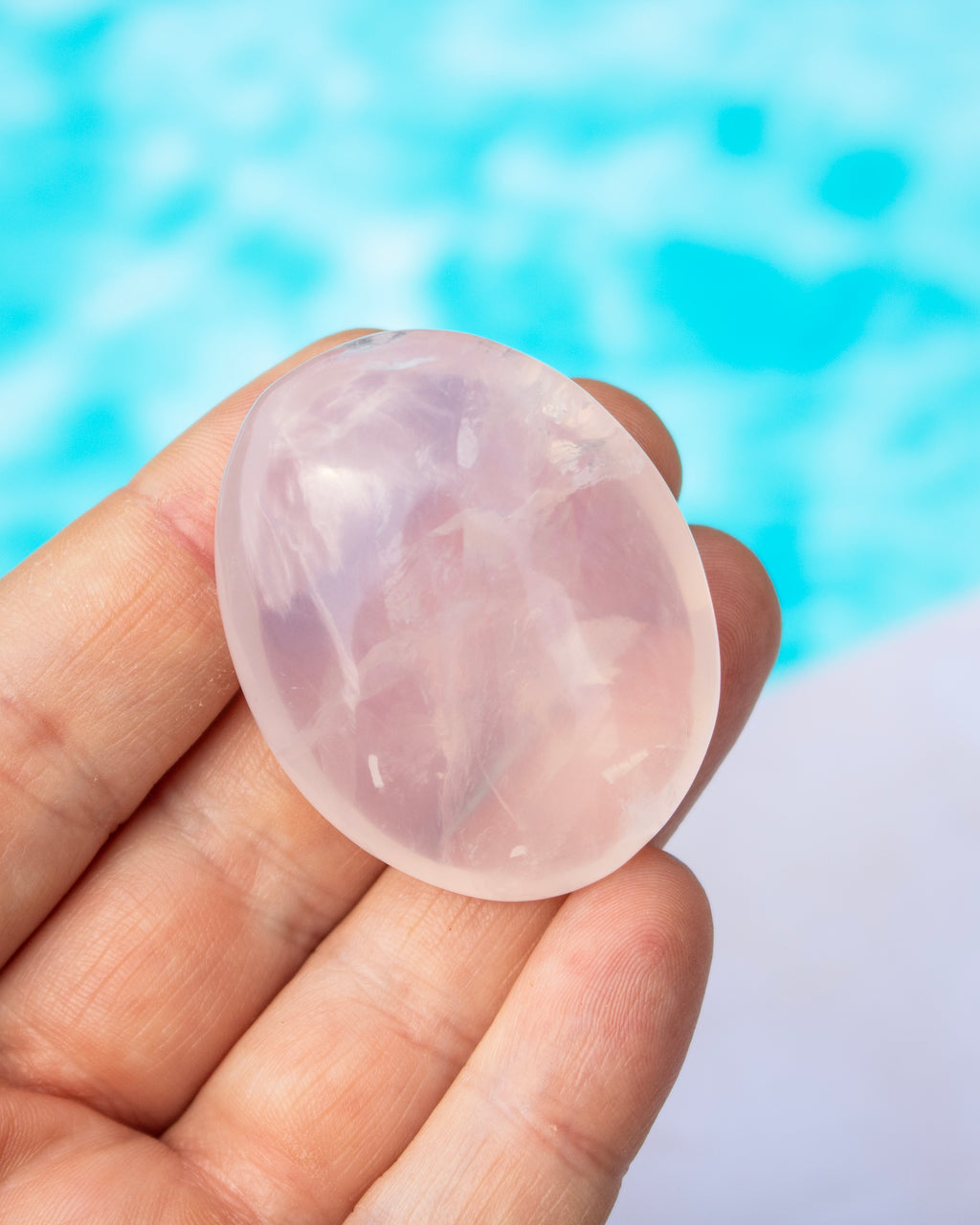 A hand holding a smooth, polished oval rose quartz gemstone against a blurred turquoise blue background.