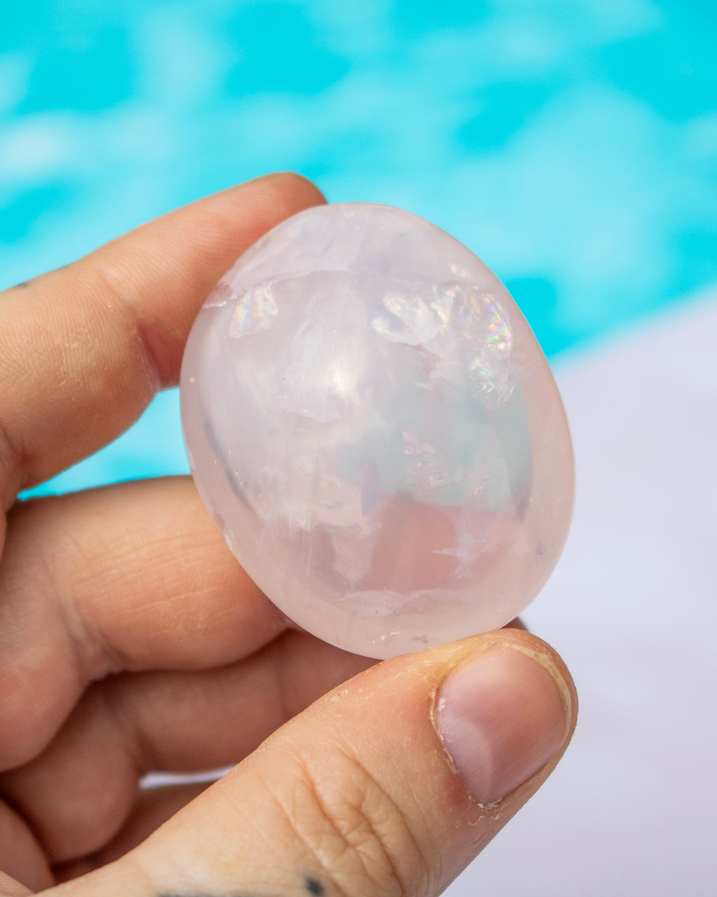 A hand holding a smooth, polished pink stone or crystal against a blurred turquoise and white background.