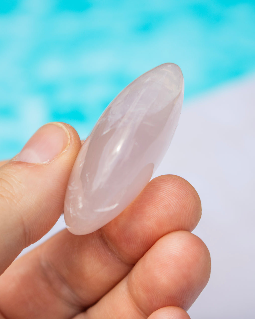 A hand holding a polished rose quartz crystal point against a blurred background of blue water and white sand.