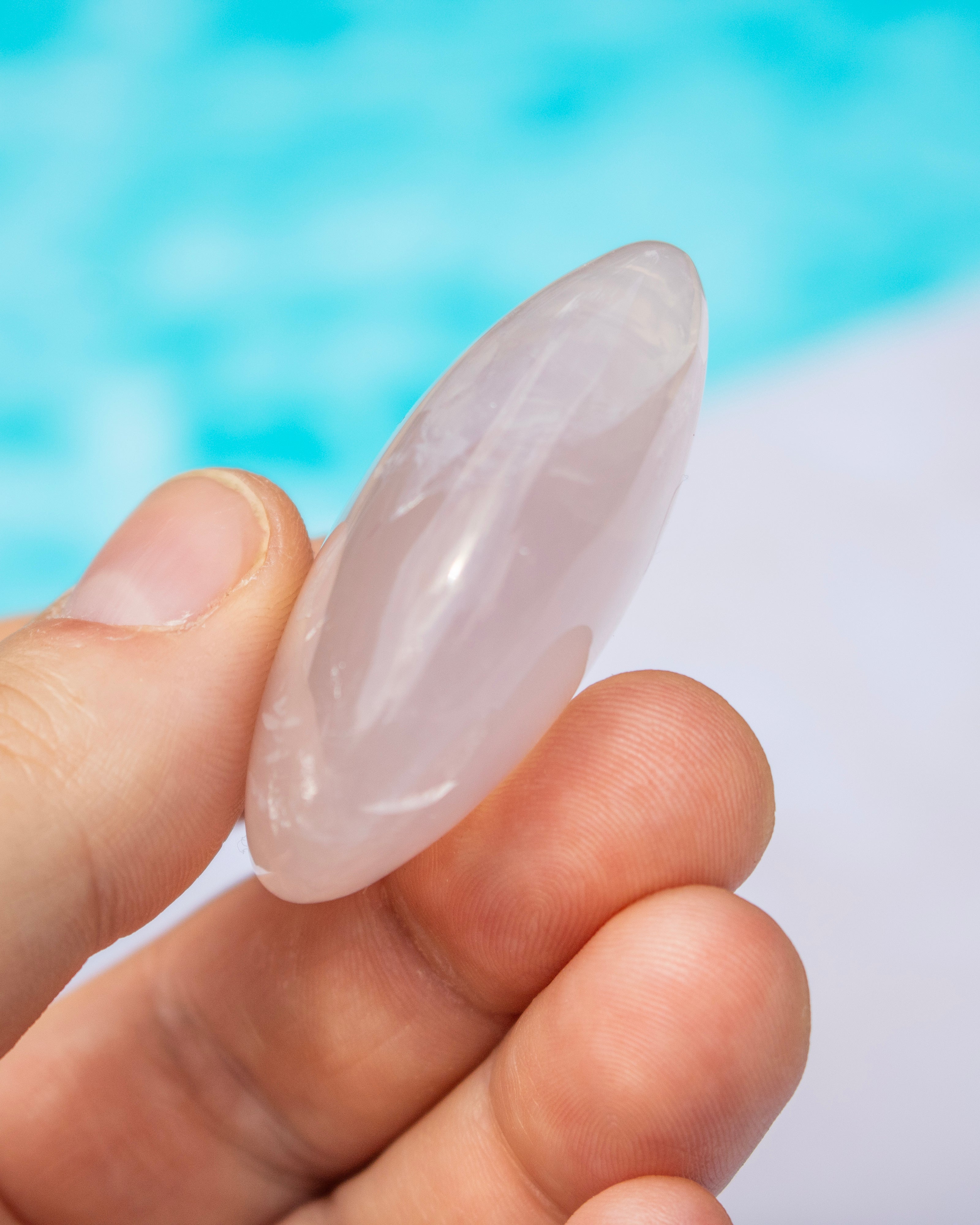 A hand holding a polished rose quartz crystal point against a blurred background of blue water and white sand.