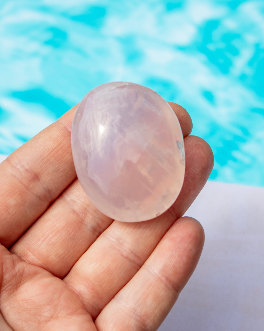 A hand holding a smooth, polished oval rose quartz crystal against a blurred background of bright blue water.