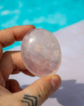 A hand holding a polished, oval-shaped pale pink crystal ball with natural inclusions, set against a blurred background of bright blue water and a white surface.
