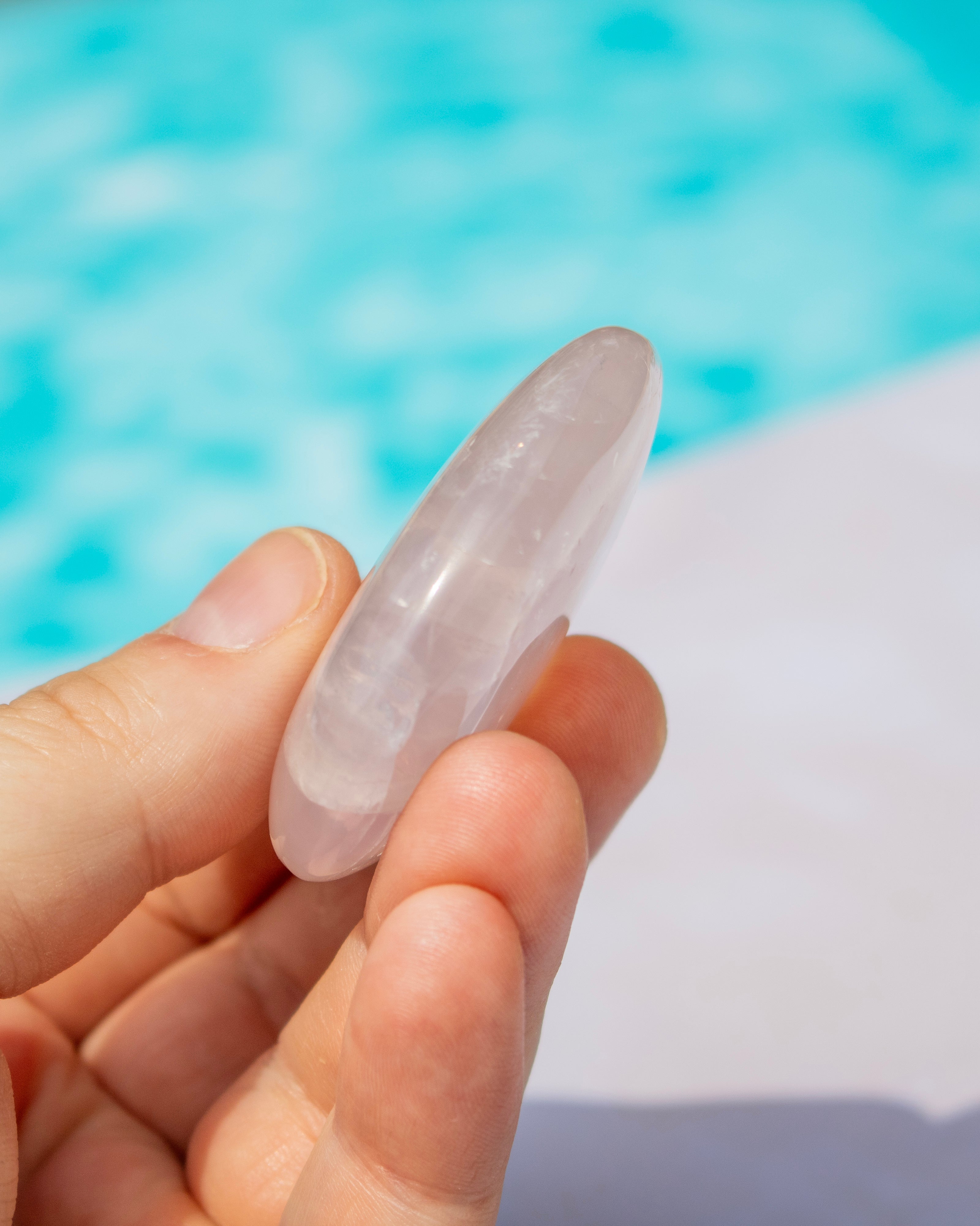 A hand holding a smooth, polished rose quartz crystal wand against a blurred background of bright blue water and a white surface.
