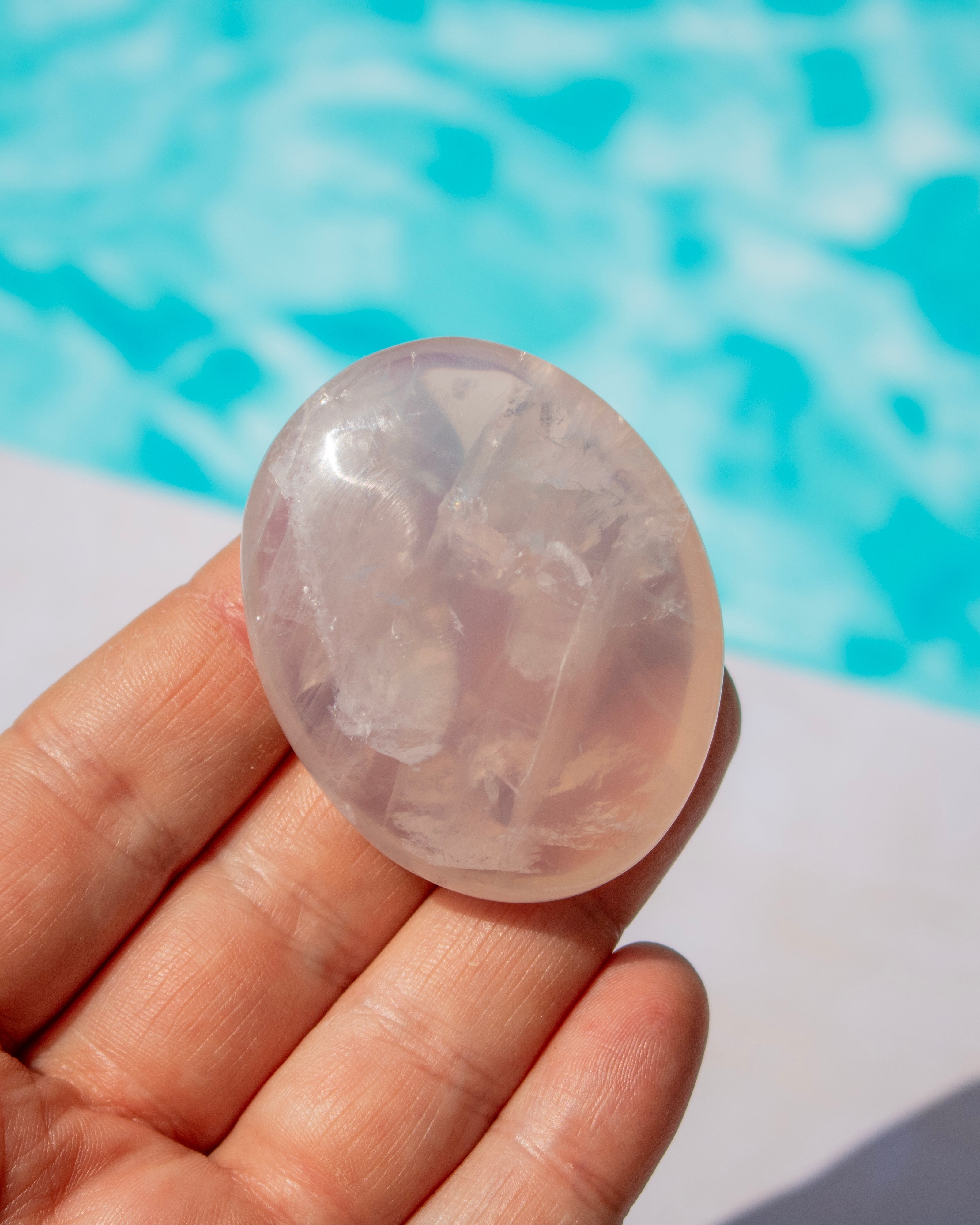 A hand holding a polished oval rose quartz crystal against a blurred background of bright blue water, likely a swimming pool.