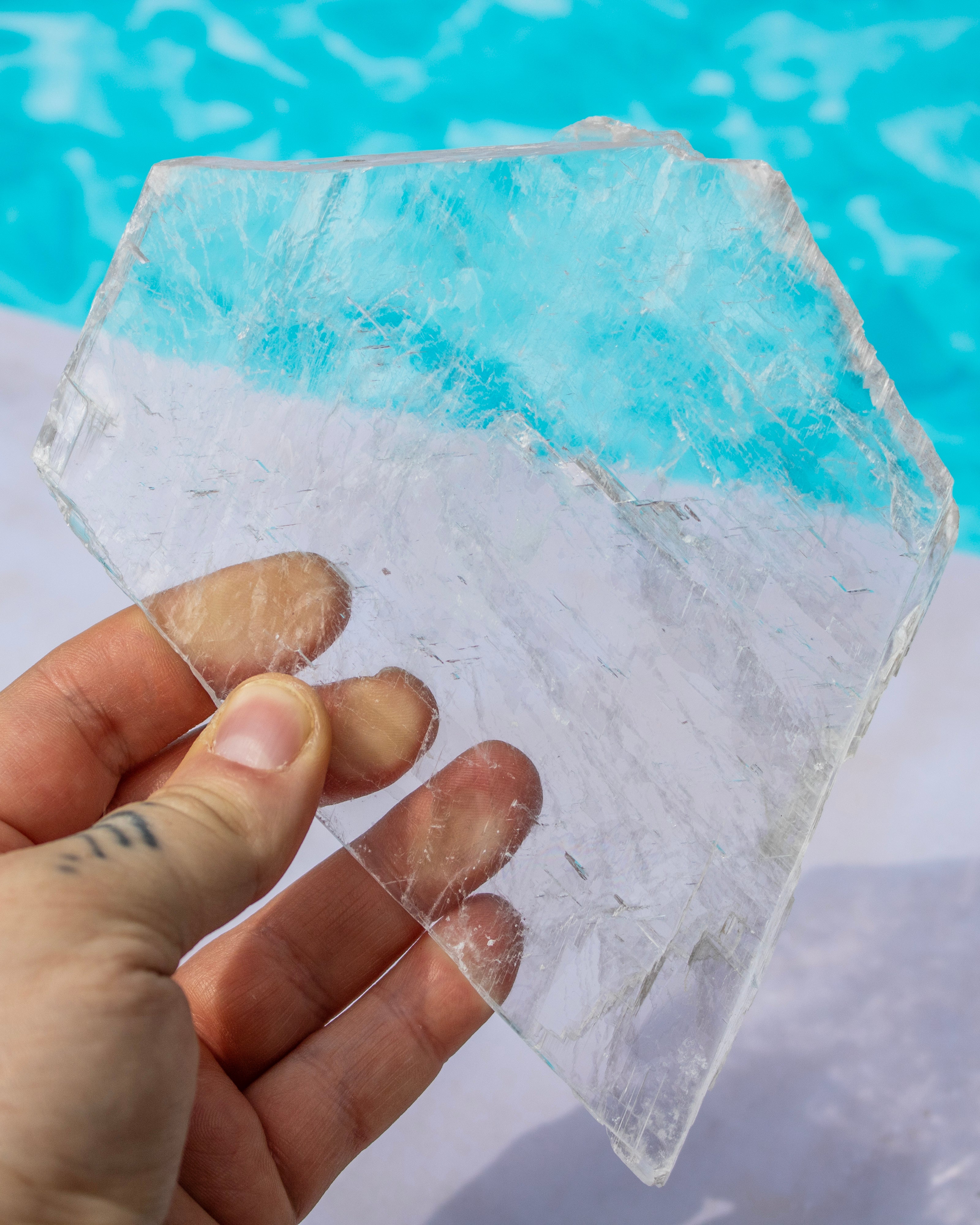 A hand holding a raw, irregularly shaped clear selenite slab with natural fracture lines, set against a background of bright blue water.