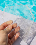 A hand holding a raw, uncut clear selenite shard in front of a bright blue swimming pool.