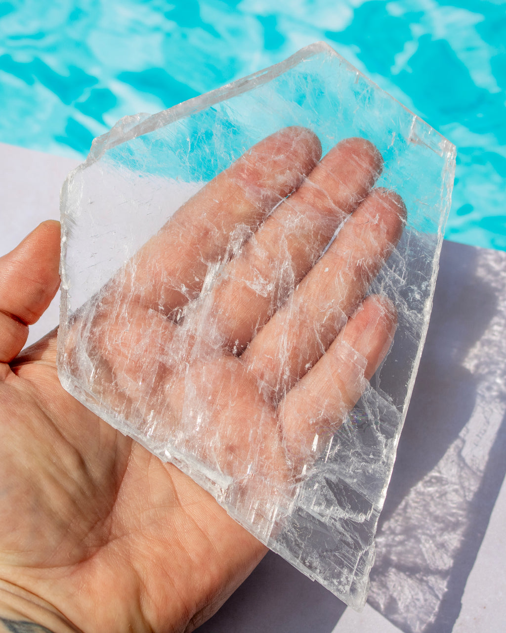 A hand holding a large, rough-cut transparent selenite outdoors with a swimming pool in the background.
