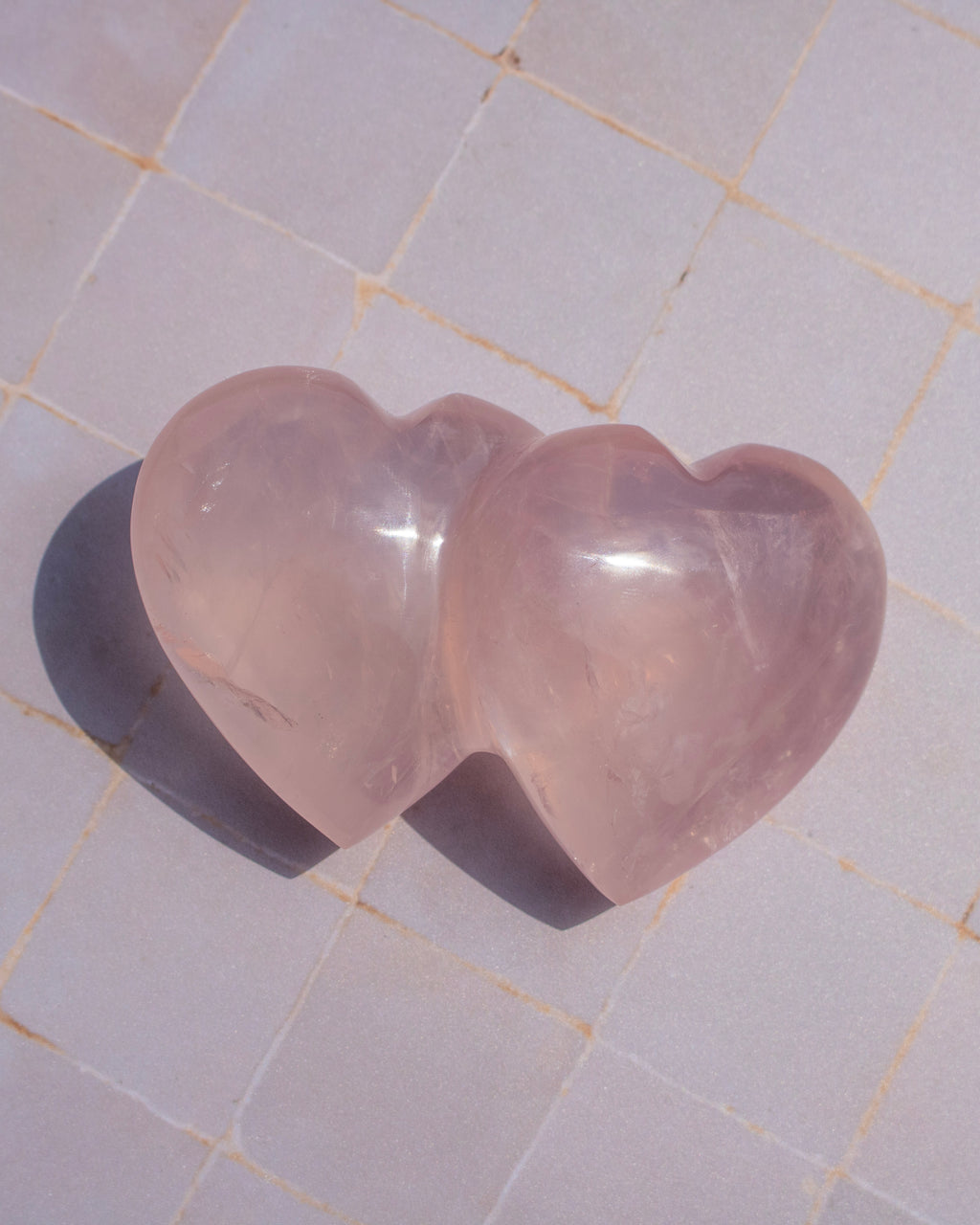 Two heart-shaped rose quartz crystal carvings resting on a light beige tiled surface, casting soft shadows.