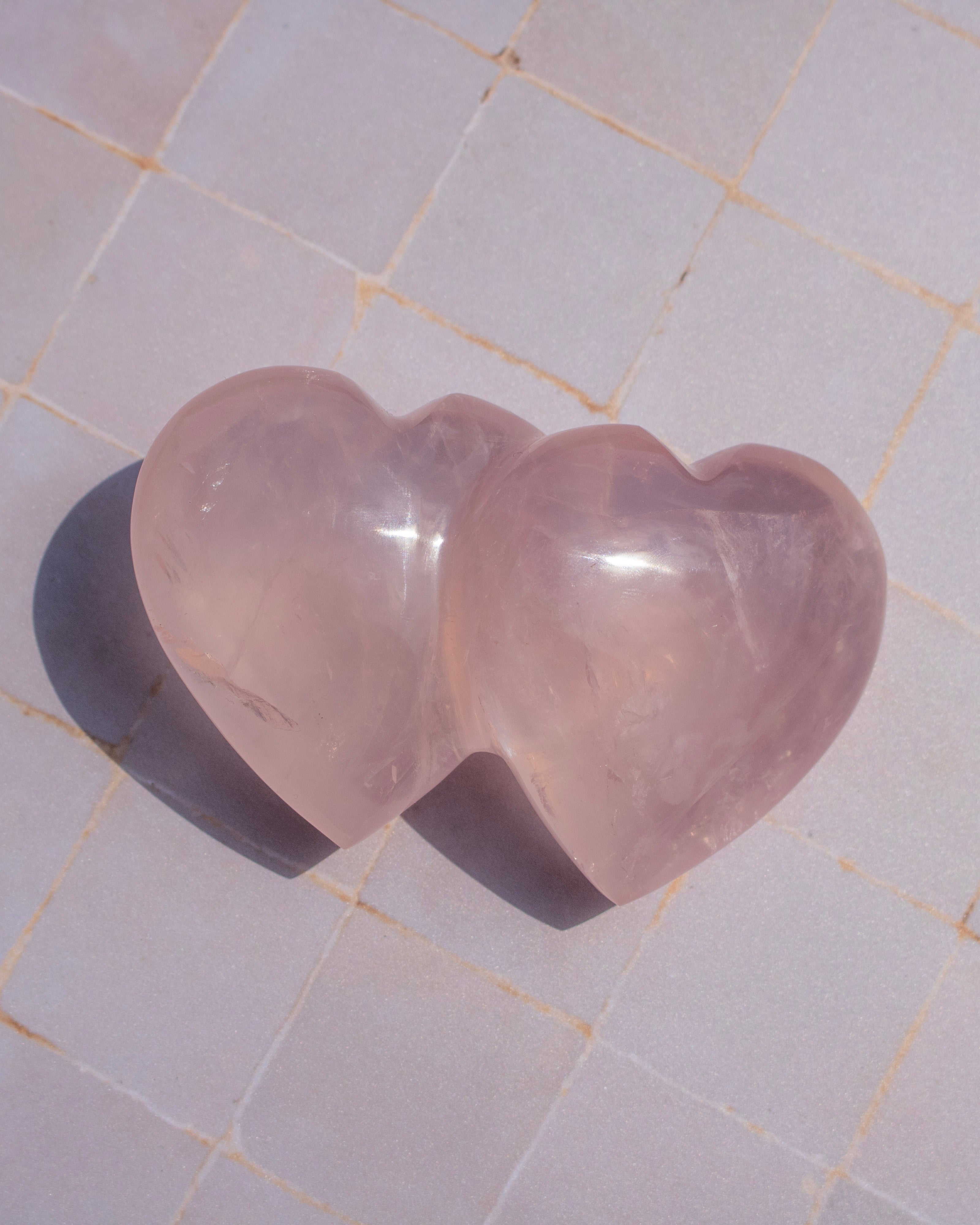 Two heart-shaped rose quartz crystal carvings resting on a light beige tiled surface, casting soft shadows.