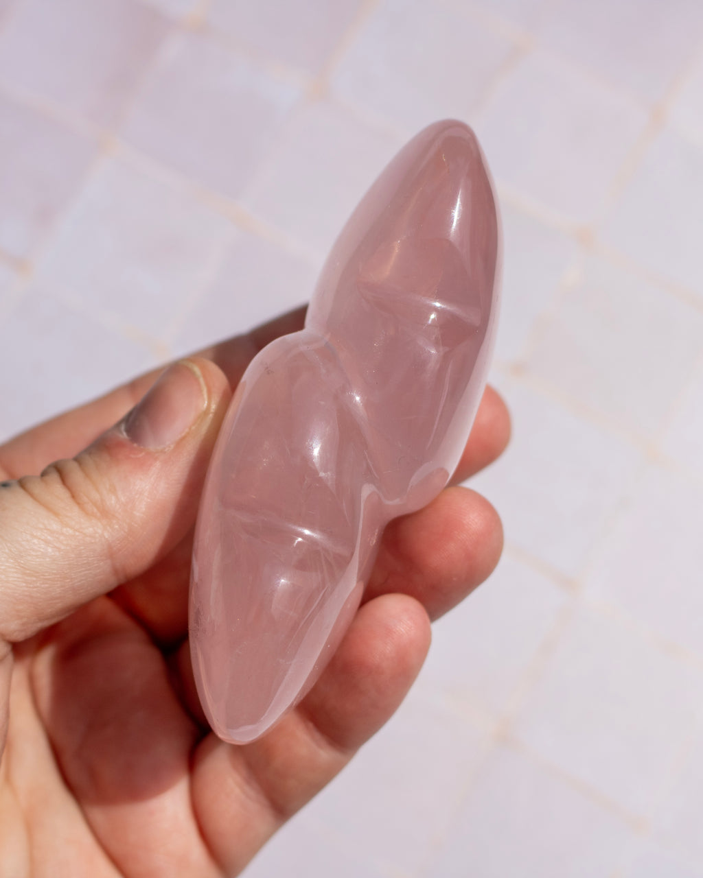 A hand holding a polished rose quartz crystal wand with a double-ended, pear-shaped design against a light tiled background.