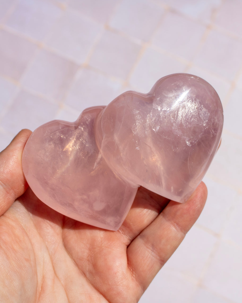 Two polished rose quartz heart-shaped crystals held in a person's hand against a light tiled background.