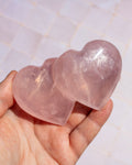 Two polished rose quartz heart-shaped crystals held in a person's hand against a light tiled background.