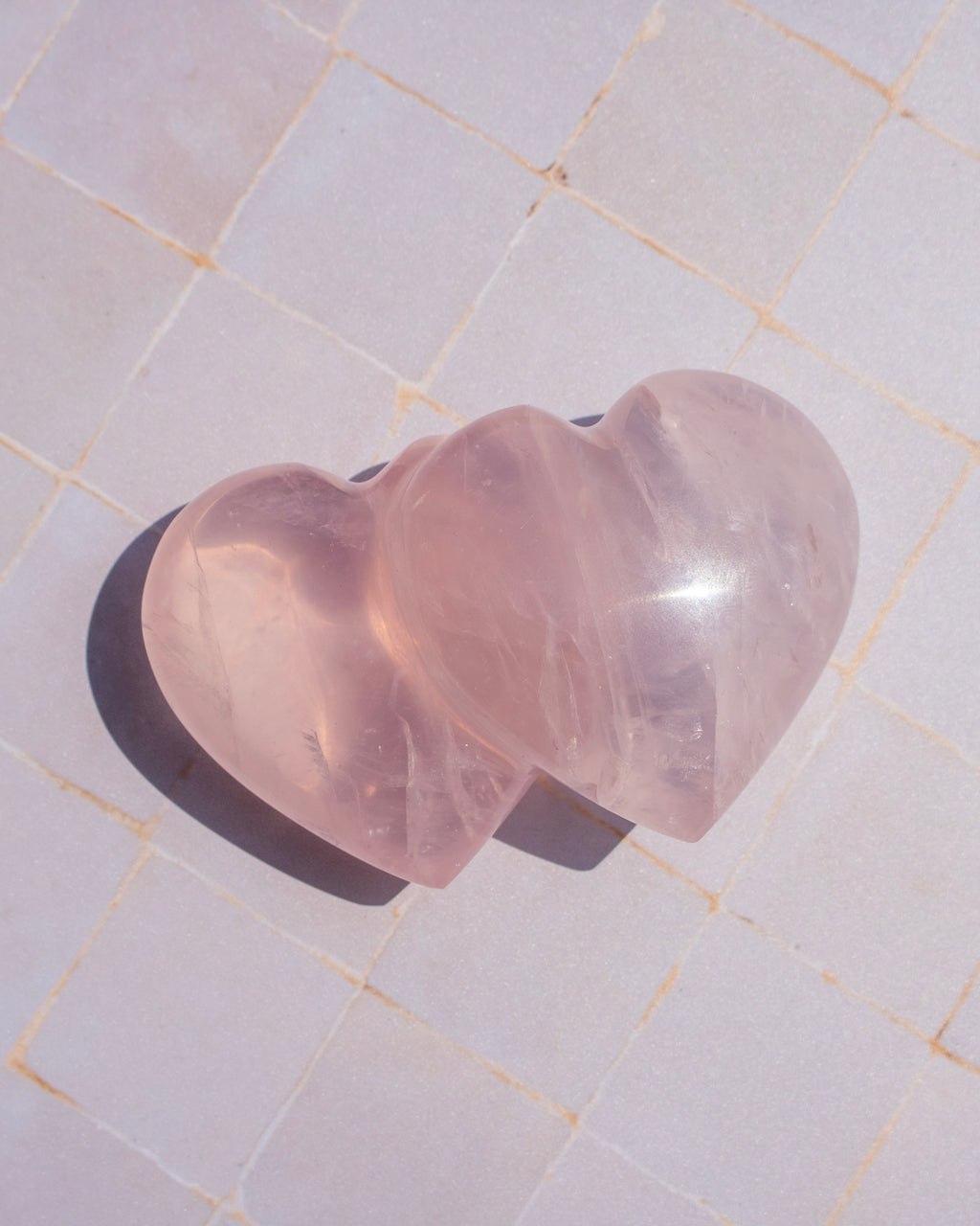 Two interlocking heart-shaped rose quartz crystal pendants resting on a light-colored tiled surface.