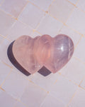 Two polished heart-shaped rose quartz crystals resting on a light beige tiled surface, casting a dark shadow to the left.