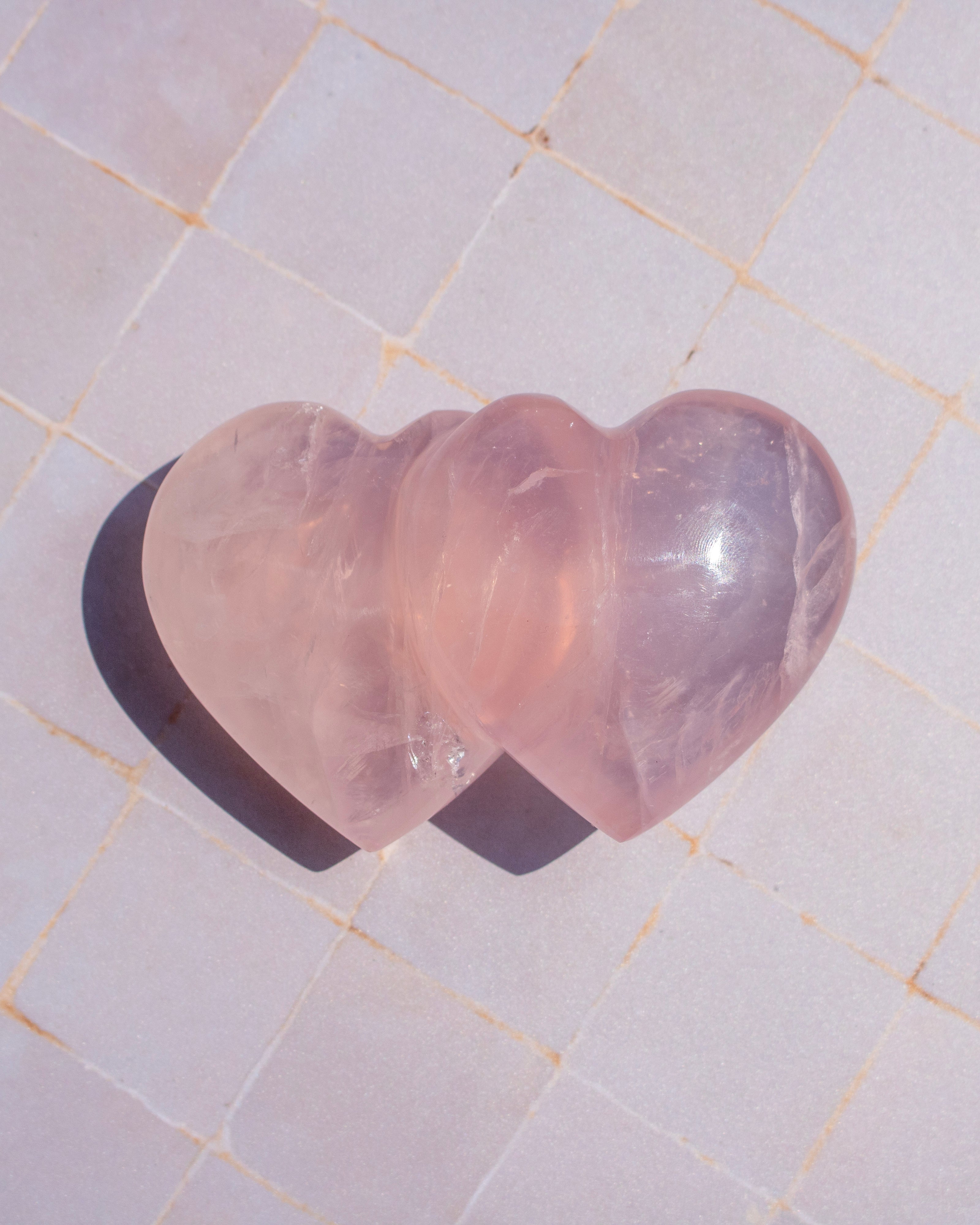 Two polished heart-shaped rose quartz crystals resting on a light beige tiled surface, casting a dark shadow to the left.