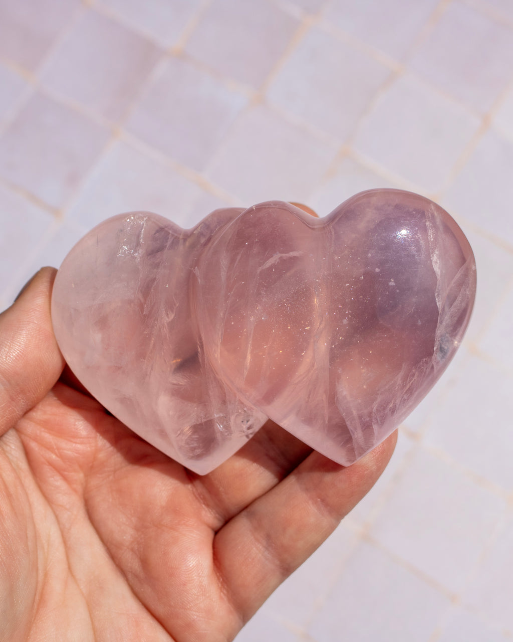 Two heart-shaped rose quartz crystal spheres held in a person's hand against a light tiled background.