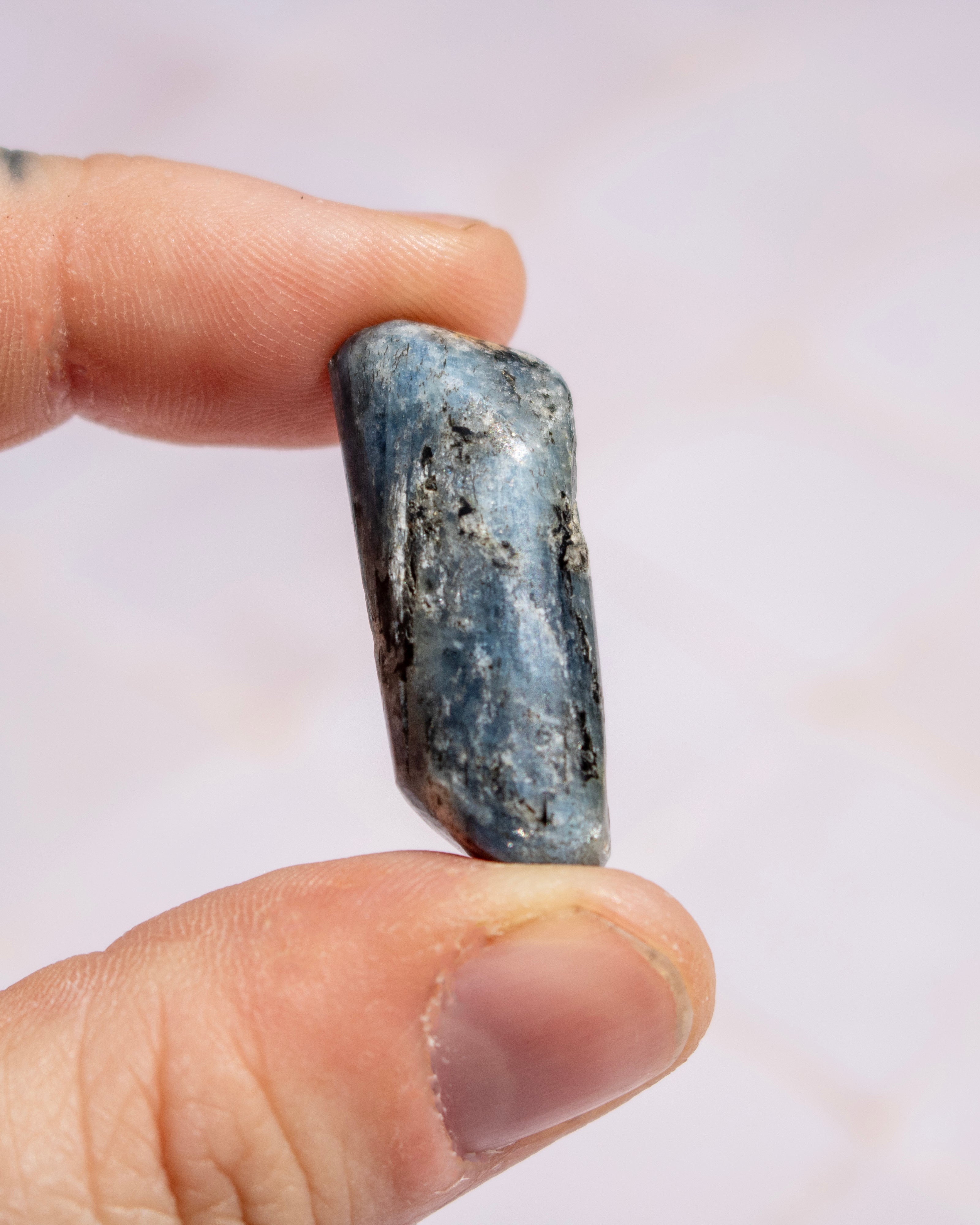 A hand holding a small polished blue-gray stone or crystal against a soft light pink background.