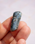 A polished blue-gray stone bead or cabochon held between fingers against a soft light background.