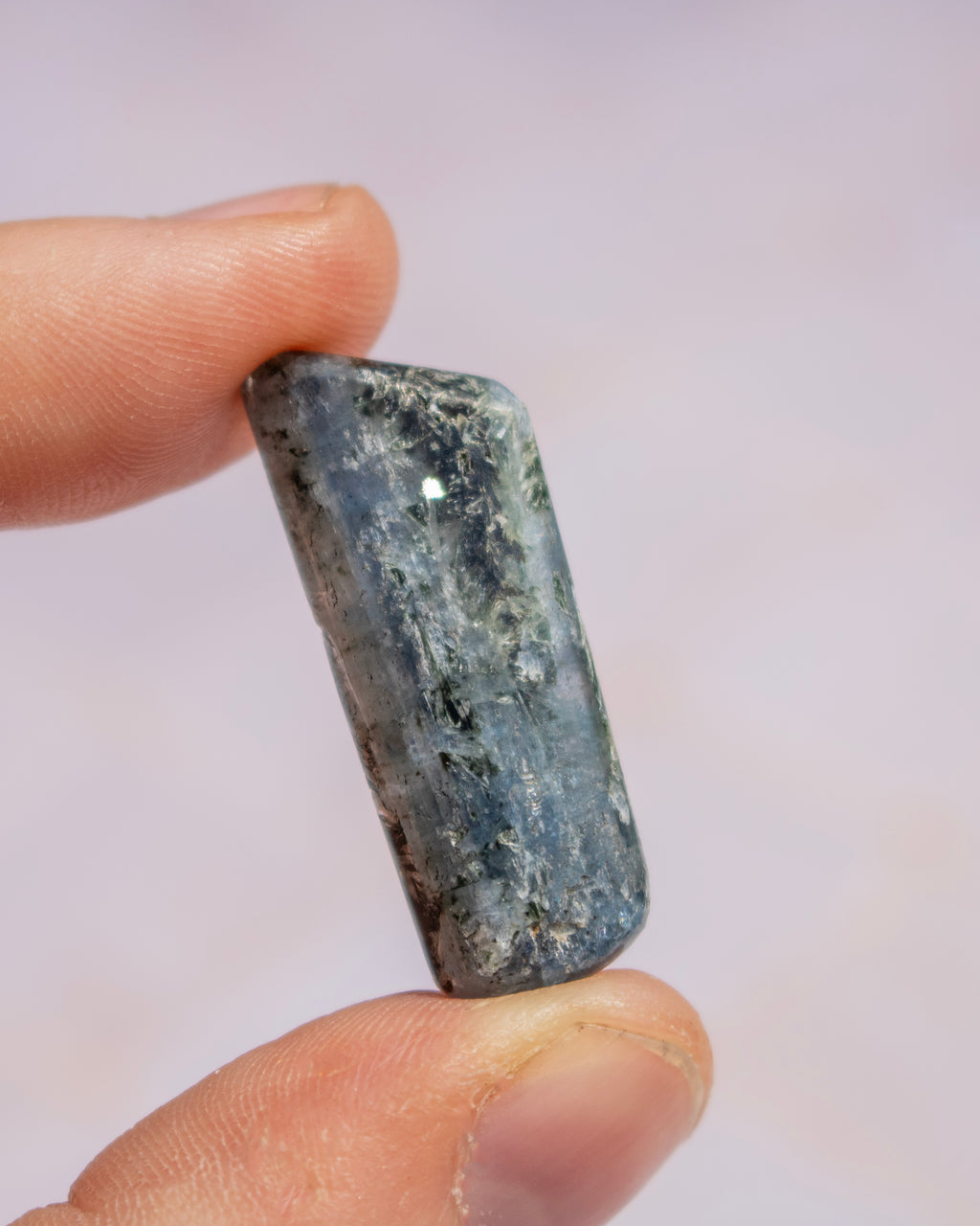 A polished rectangular stone with a mottled slate gray and blue-green coloration, held between two human fingers against a soft, light pink background.