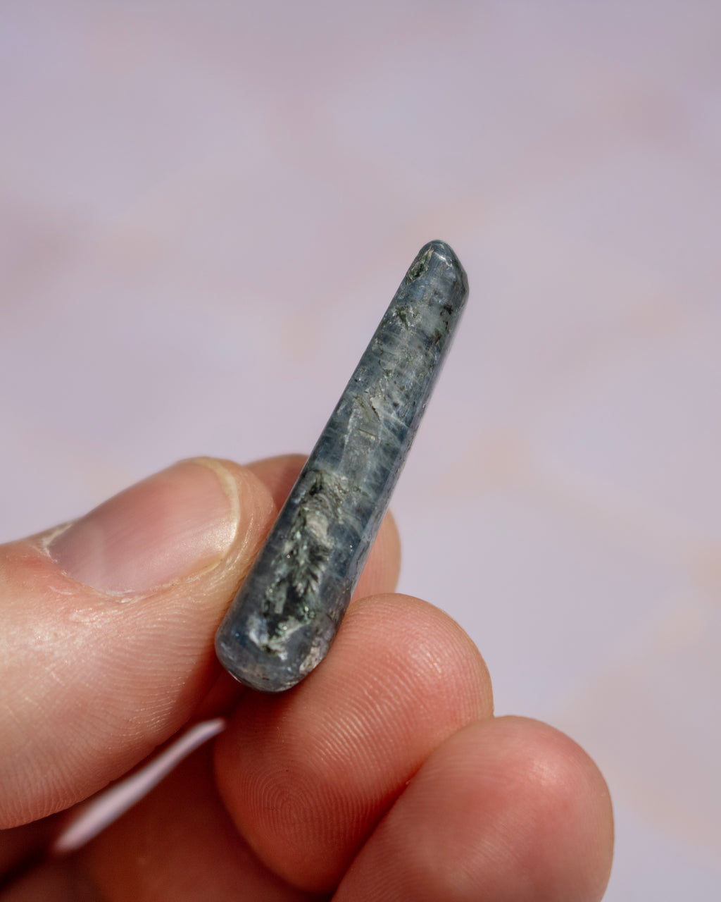A small polished gray stone or crystal pointed tip held between fingers against a soft light pink background.