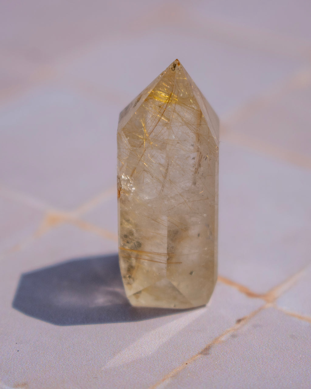 A natural clear quartz point crystal with prominent golden needle inclusions, standing upright on a light marble surface.